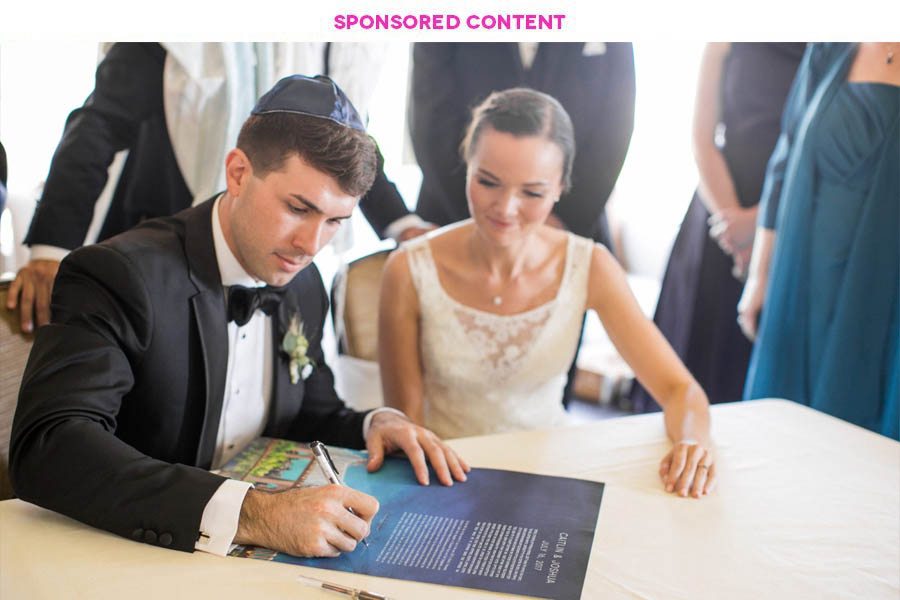 man in tux and yarmulke signs a beautifully designed ketubah as his new wife sits beside him