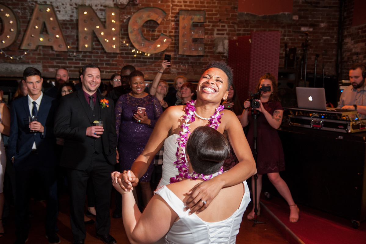 Black bride wearing a lei grinning and dancing with her wife on the dance floor while guests look on  in a photo by Leise Jones
