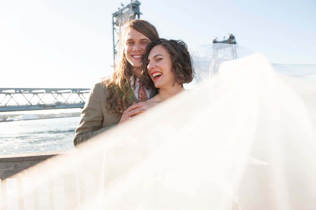 Groom with long hair and bride with short hair and red lipstick stand with their backs to a waterfront laughing while the bride's veil swirls in front of them in a photo by Leise Jones