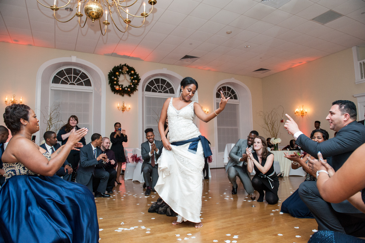 An Indian bride in a white dress dances in the center of a ballroom as guests kneel around her clapping  in a photo by Leise Jones