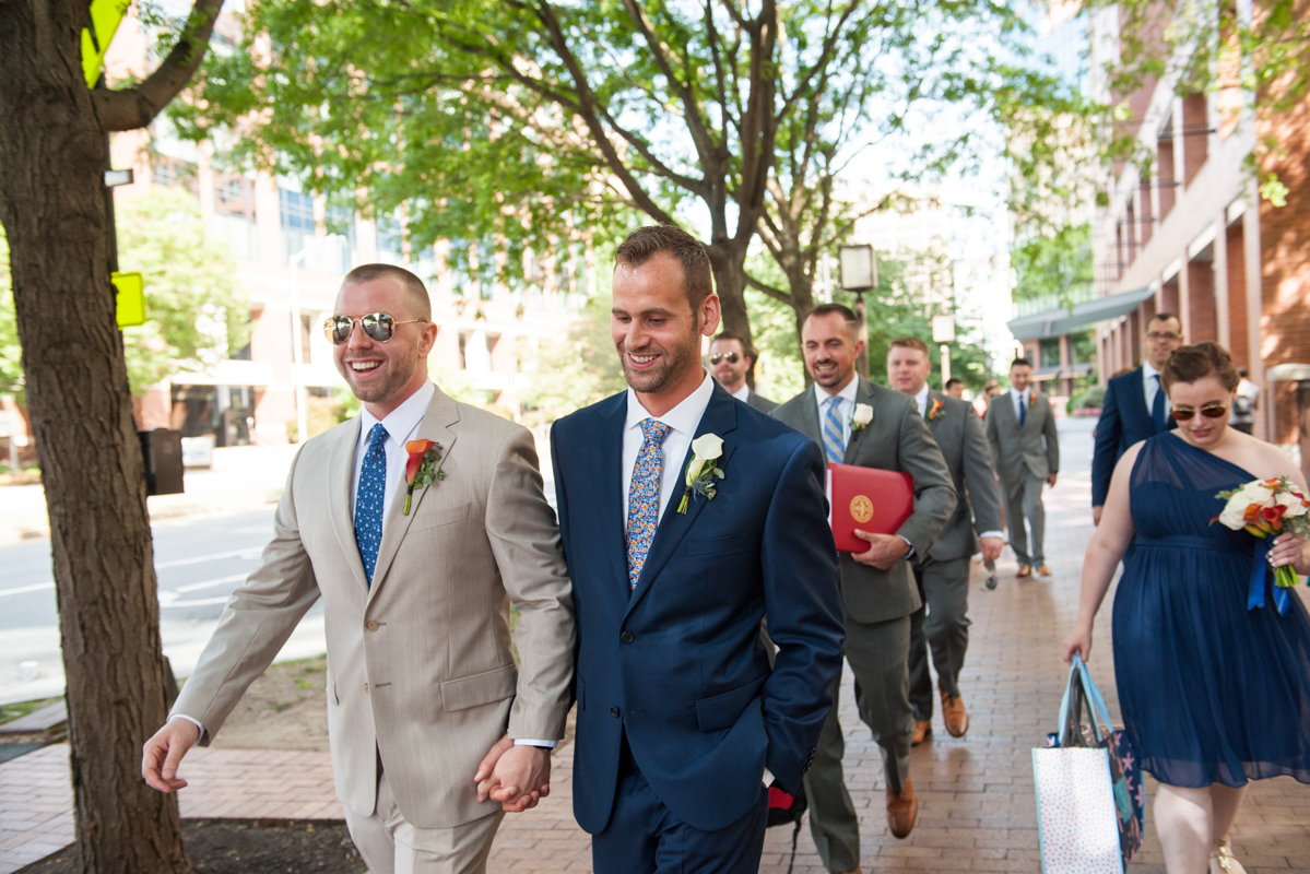 Two grooms walk down a tree-lined city street holding hands and smiling while their wedding party of men in grey suits and women in navy dresses walks behind them  in a photo by Leise Jones