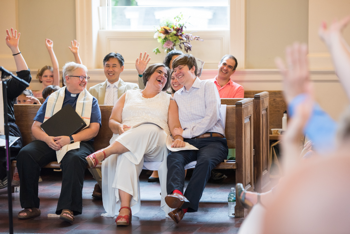Queer wedding couple are seated in the front pew of a chapel next to their minister, and lean towards each laughing and holding hands  in a photo by Leise Jones