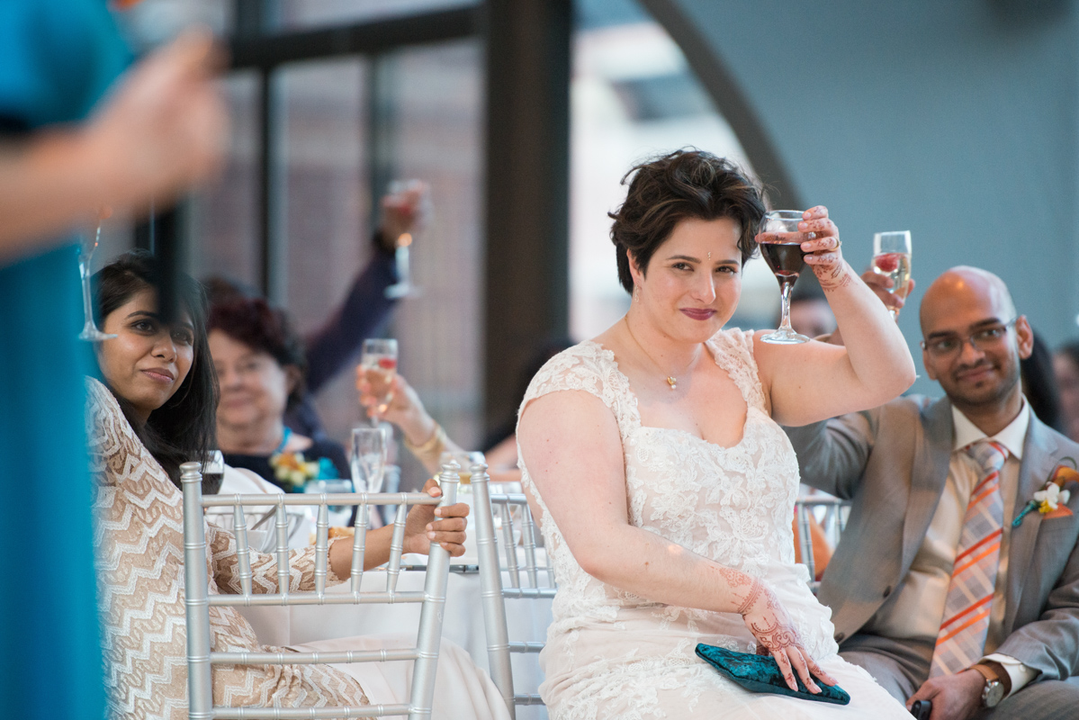 Bride with short dark hair is seated at the dinner table and lifts her wine glass in a toast towards a speaker who is not shown  in a photo by Leise Jones