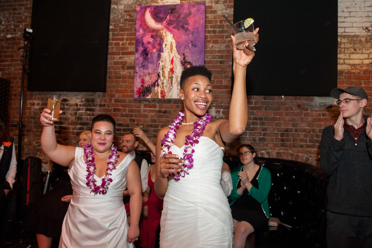 Two brides in white gowns and purple leis lift glasses to toast while standing in an industrial looking reception space with exposed brick walls and modern art  in a photo by Leise Jones