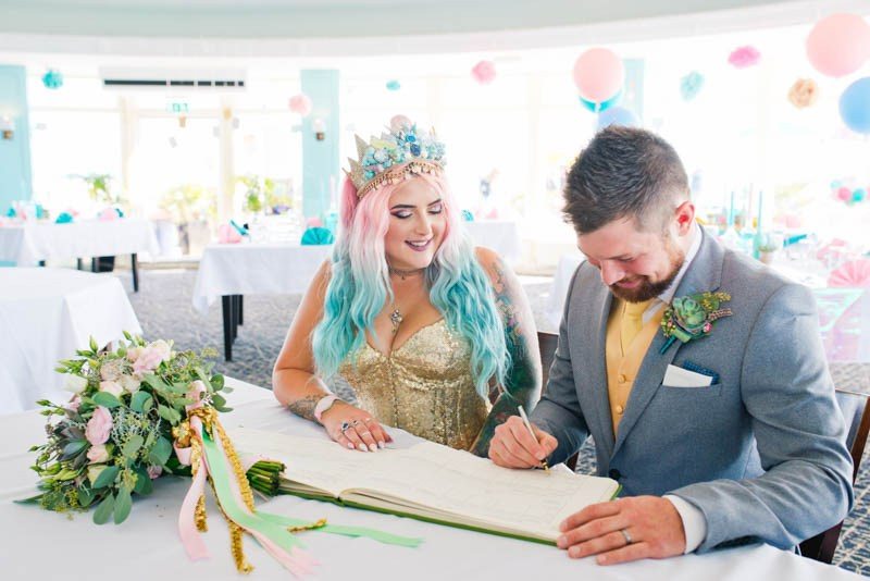 bride with pink and blue hair and a seashell crown sitting next to man signing book