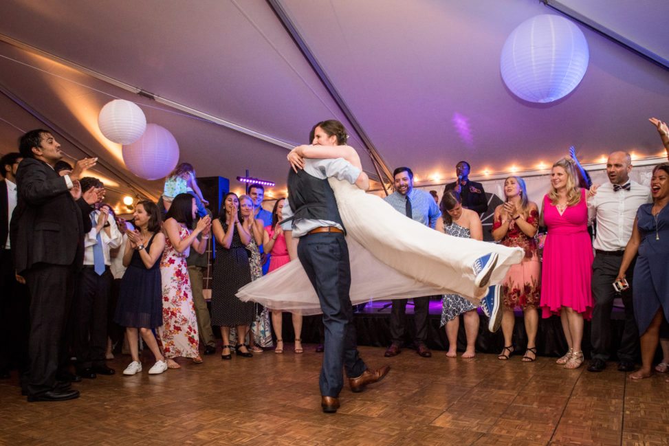 groom swings bride around the dance floor of a wedding tent so her white dress flows out in a circle around them as guests make a circle around them and cheer them on  in a photo by Leise Jones