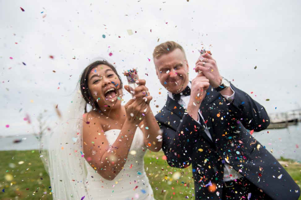 Laughing Asian bride with white dress and veil and red-headed white groom in a back suit explode poppers so colorful confetti floats all around them in a photo by Leise Jones
