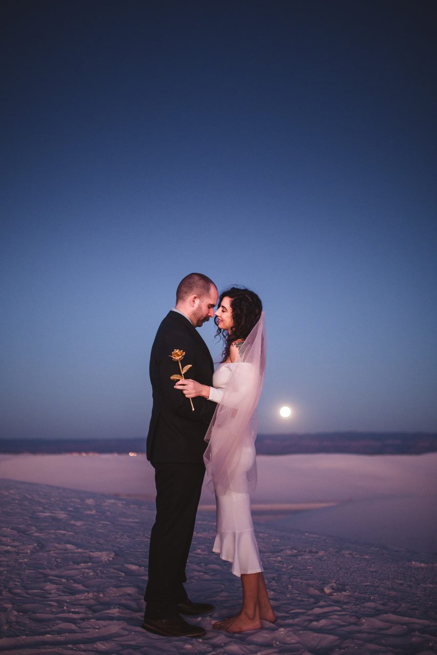 A couple stands together in the moonlight on white sands