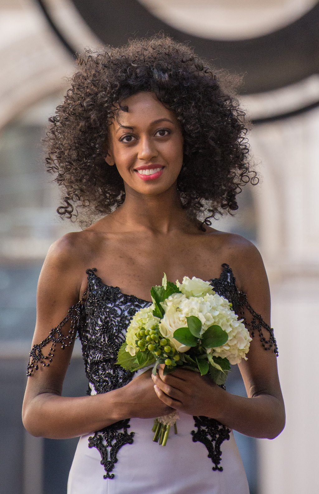 a woman in a white wedding gown with lacy beaded black overlay smiles at the camera while holding a bouquet