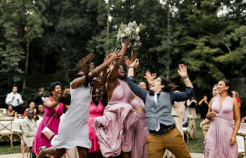 a group of people in wedding guest finery jump and reach for a bouquet