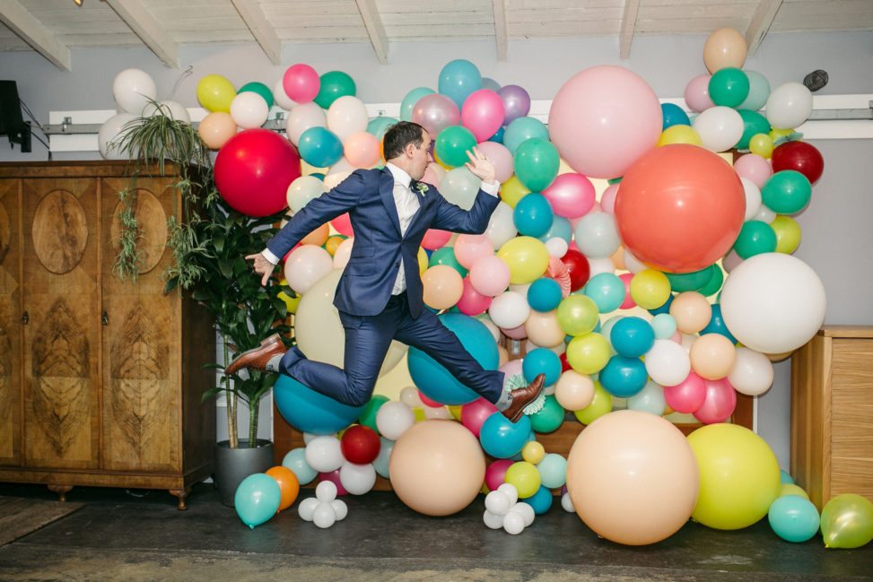 man jumps into air in front of wall of balloons