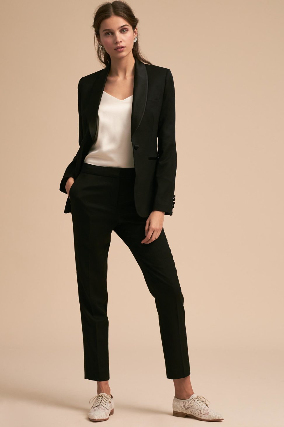 A woman stands, dressed in a black tuxedo jacket and black pants with white shoes.