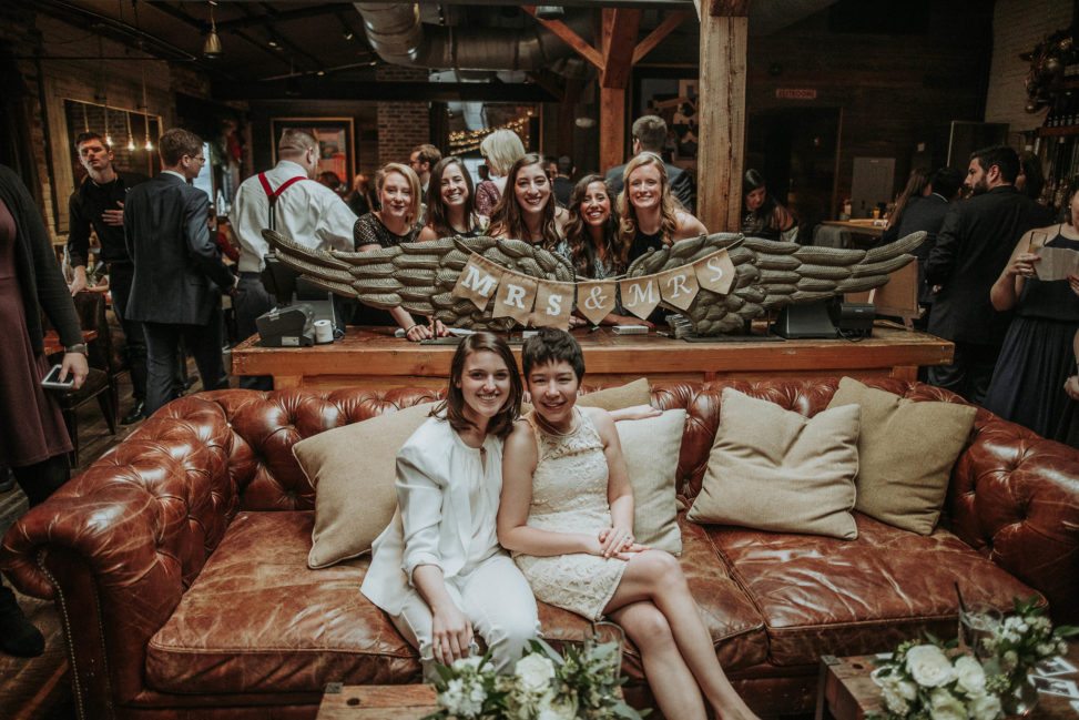 two brides sitting on leather couch in front of friends and "Mrs. and Mrs." bunting sign