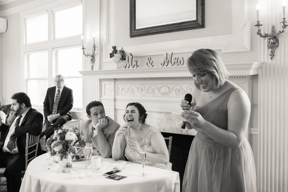 black and white image of bride and groom sitting at table and laughing during MOH speech
