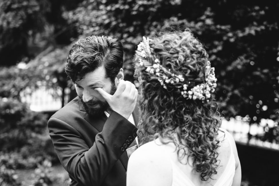 black and white image of groom wiping a tear from his eye while looking at bride with floral crown