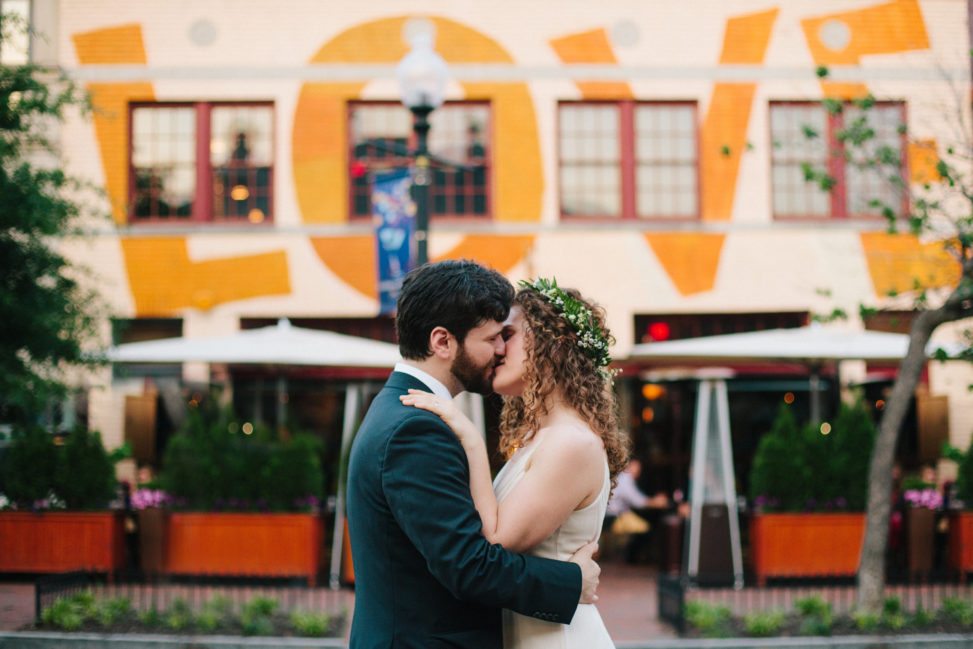 bride with flower crown and groom kissing in front of building with "Love" written on it