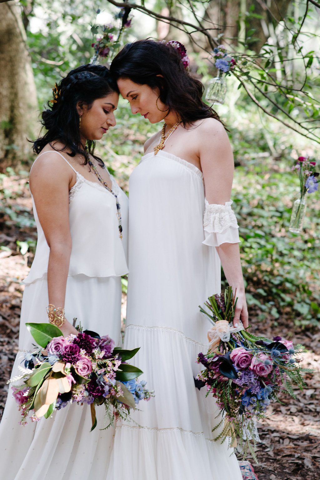 two brides touch foreheads while standing in a forest holding large, natural purple, pink, and blue bouquets