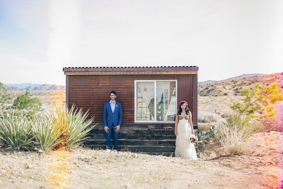 bride and groom pose separately outside a small shack in a desert