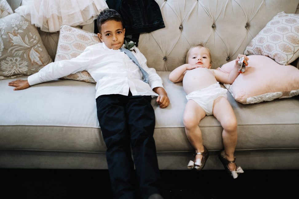 a young child in a tie and untucked shirt and a baby in a diaper and silver bowed mary janes lie on a couch looking spent