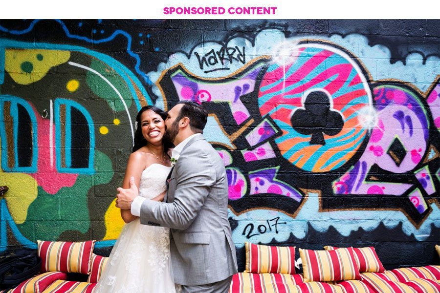 Groom in grey suit kisses bride in white dress in front of brightly colored wall with graffiti lettering