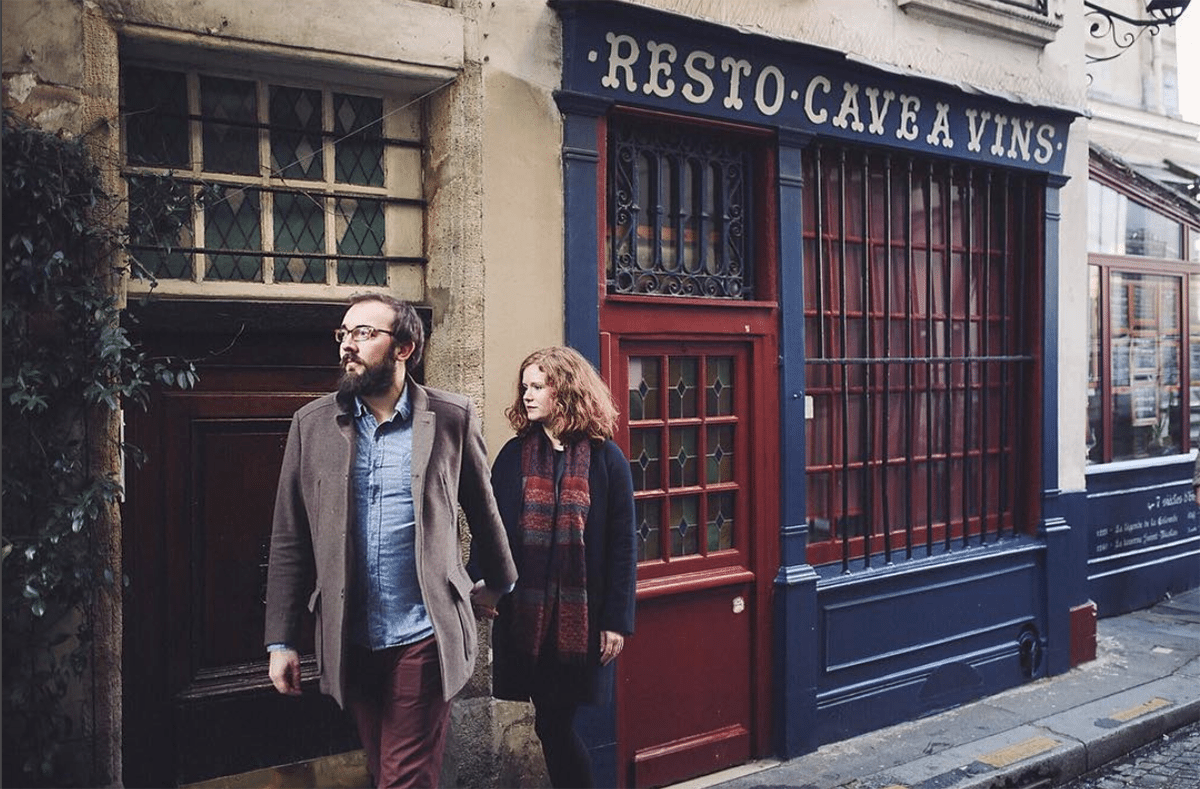 two people hold hands while walking down a street and not looking at the same things—the blues and reds of their clothes coordinate with the building they are standing in front of, which has a sign that reads "Resto-Cave a Vins"