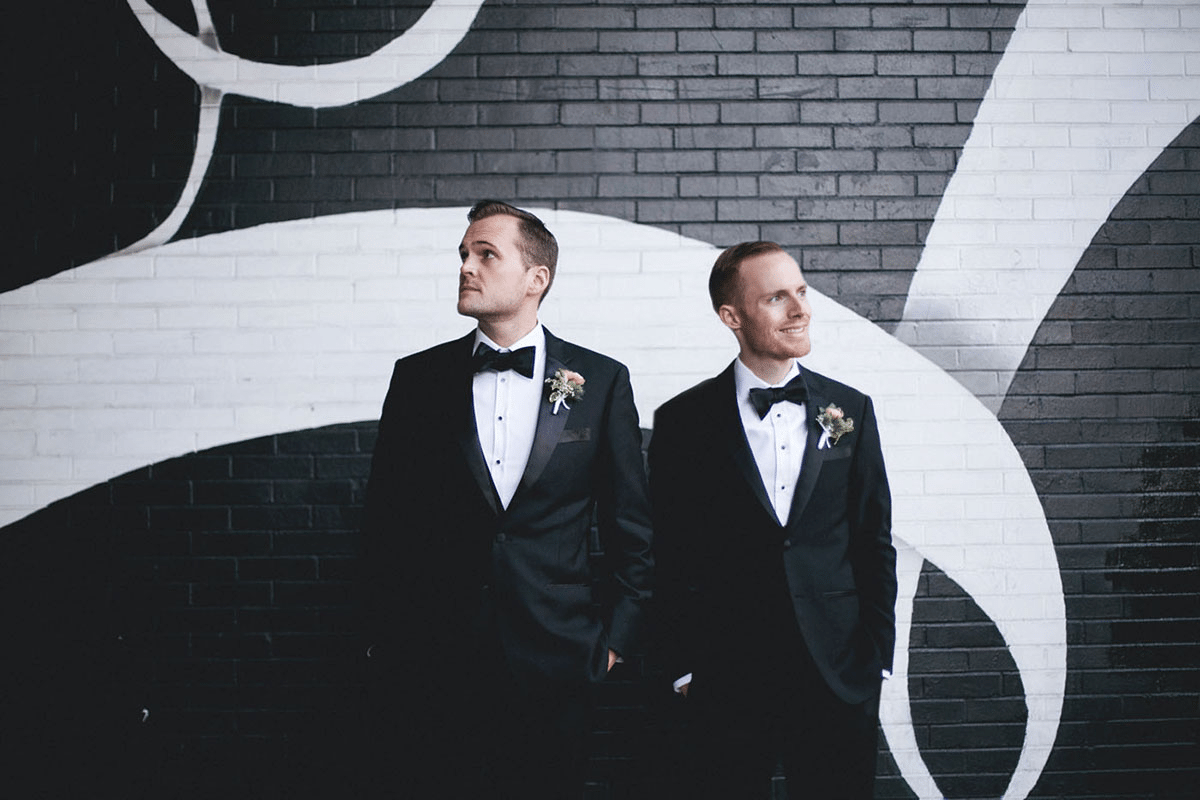 two men stand beside each other wearing tuxes looking in opposite directions, with one man smiling and the other man questioning life
