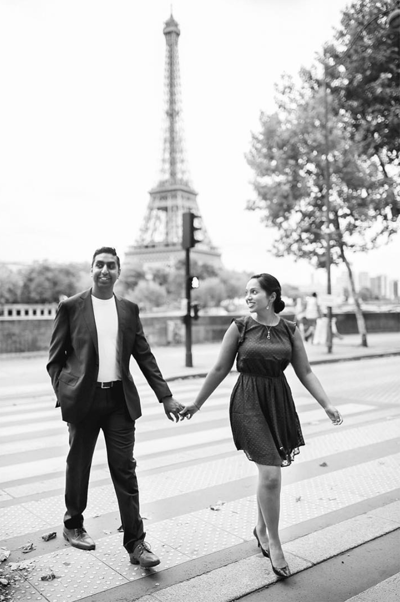 a couple walk through a park, holding hands and smiling, with the Eiffel tower behind them