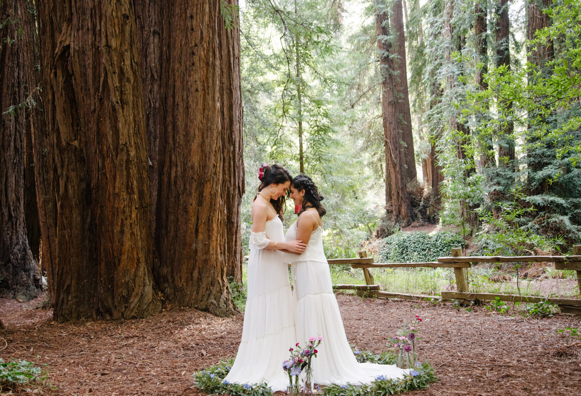 two women in white dresses touch foreheads in the woods