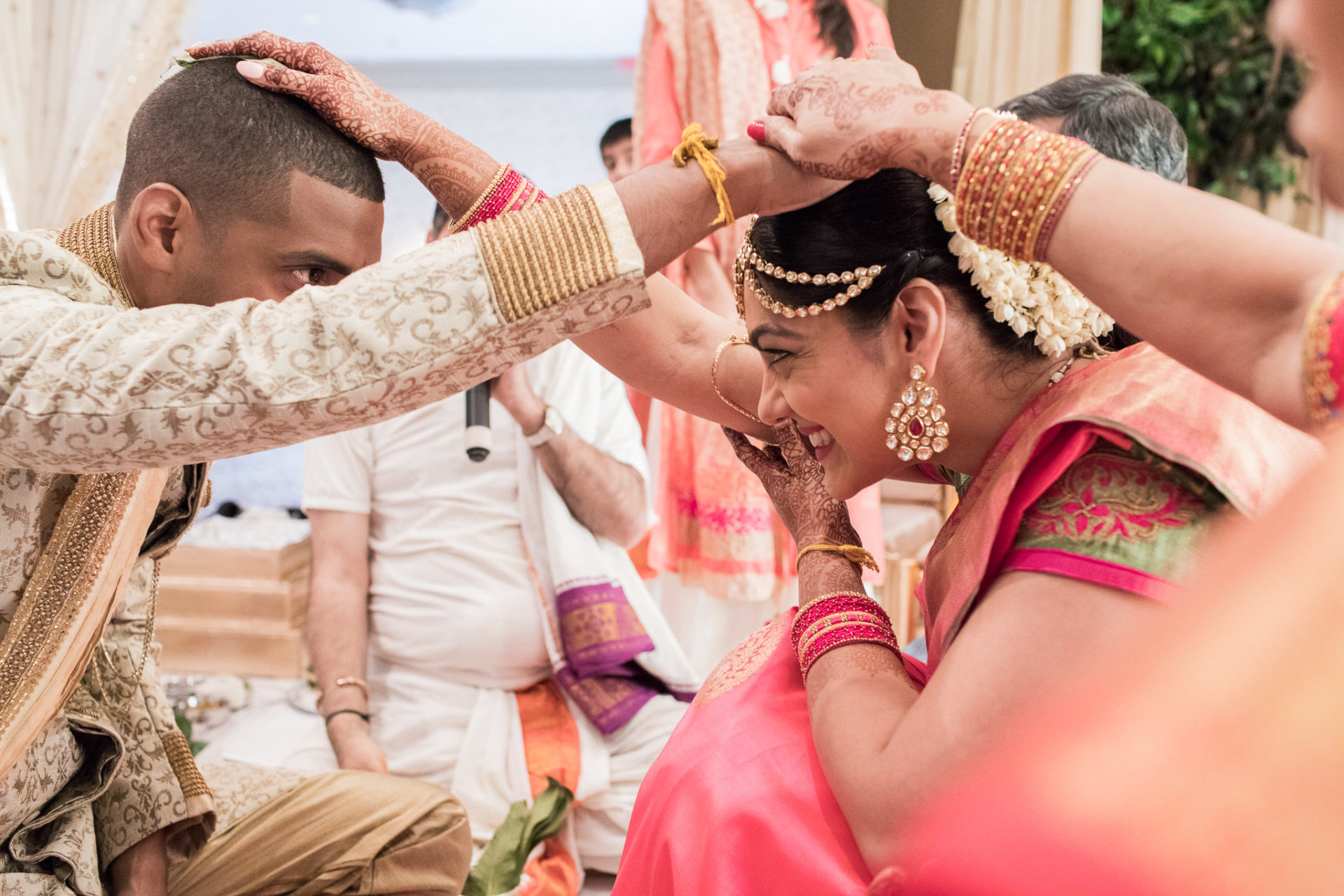 a woman with mendhi rests her hand on the grooms hand, which lays on the bride's head. the bride rests her hand on the grooms head, smiling, wearing a bright pink sari.