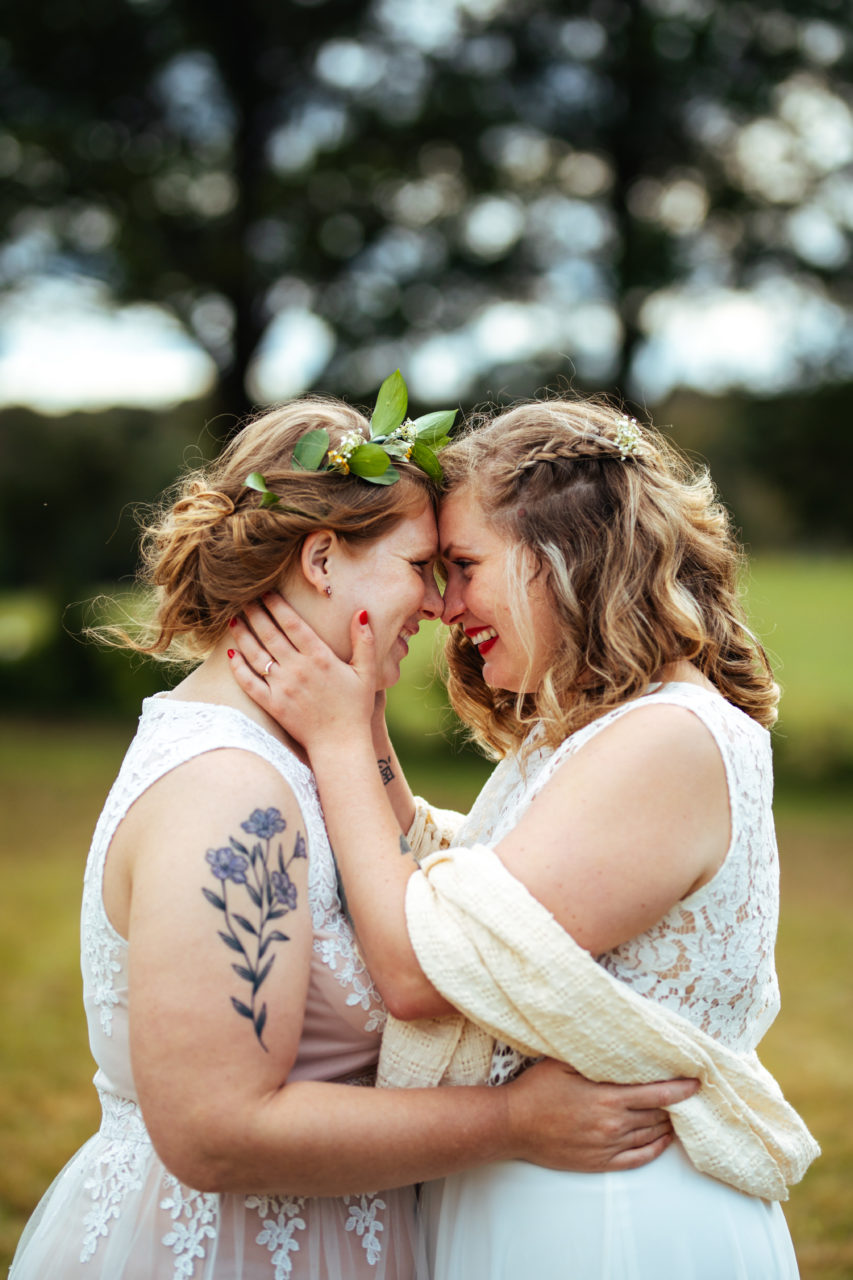 Two women in wedding dresses smile at each other