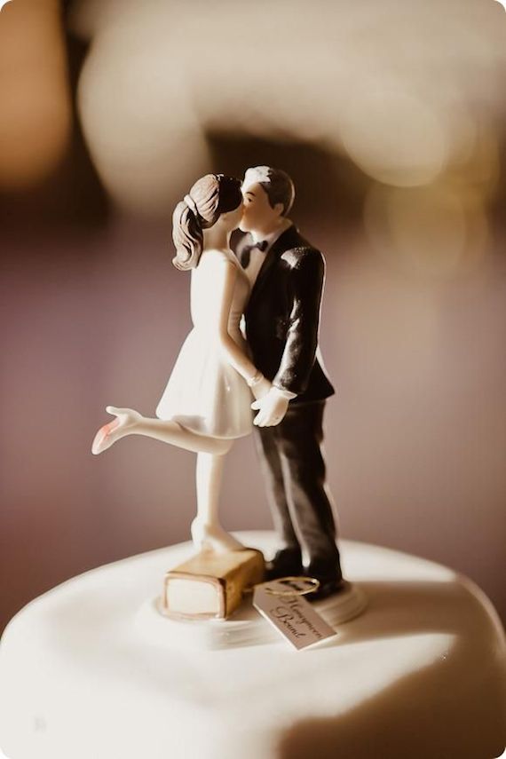 bride in a short dress and groom kissing wedding cake topper, the bride stands on a suitcase with a tag that reads "honeymoon bound"