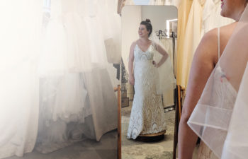 Woman in a bridal salon tries on a wedding dress, smiling, looking away from the mirror she is reflected in