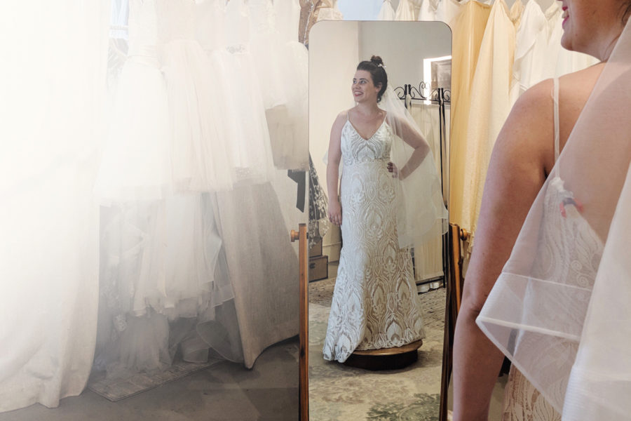 Woman in a bridal salon tries on a wedding dress, smiling, looking away from the mirror she is reflected in