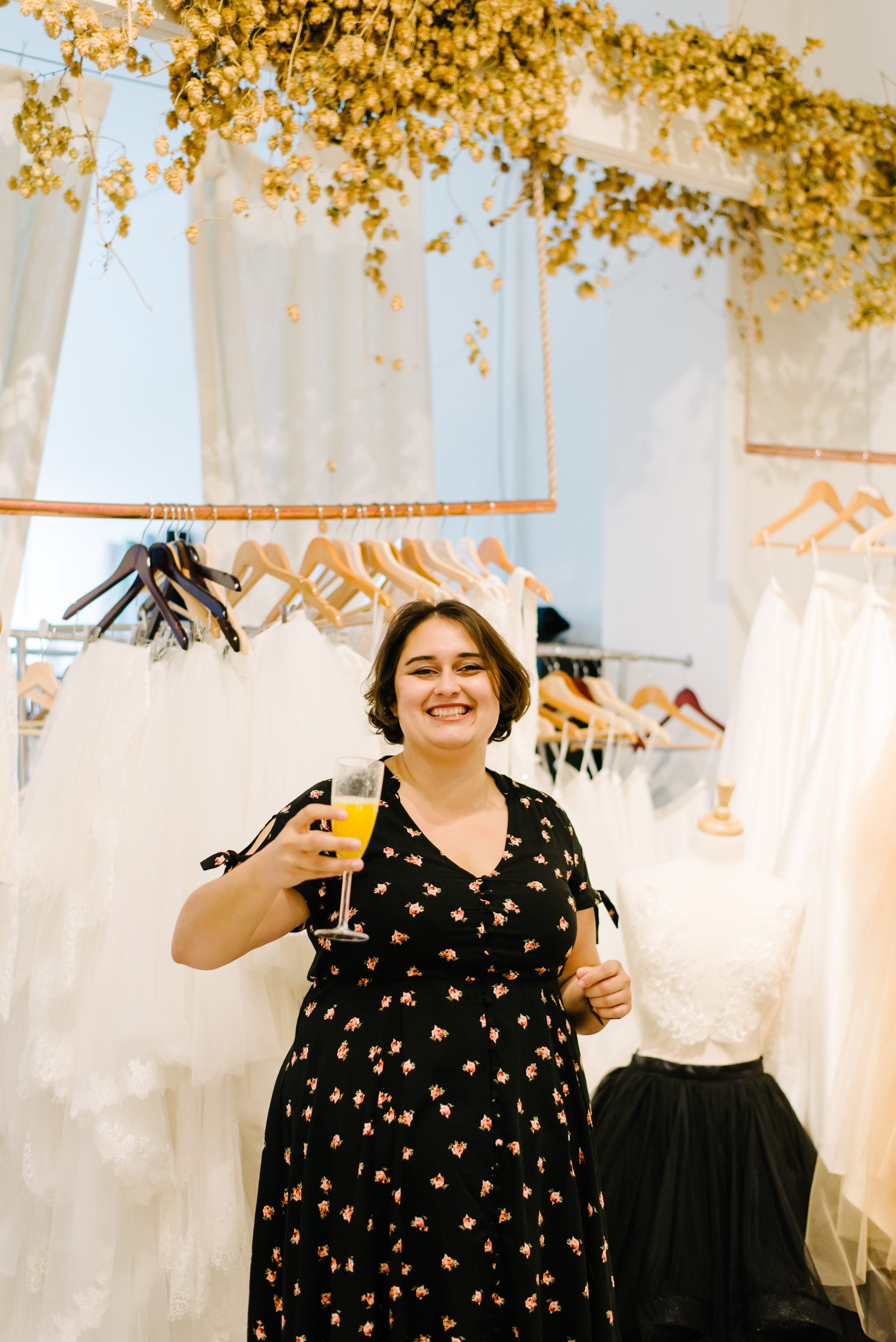 a woman holds a mimosa towards the camera while standing in front of many wedding dresses