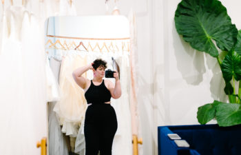 A beautiful woman wearing all black, stands and adjust her hair while surrounded by wedding dresses.