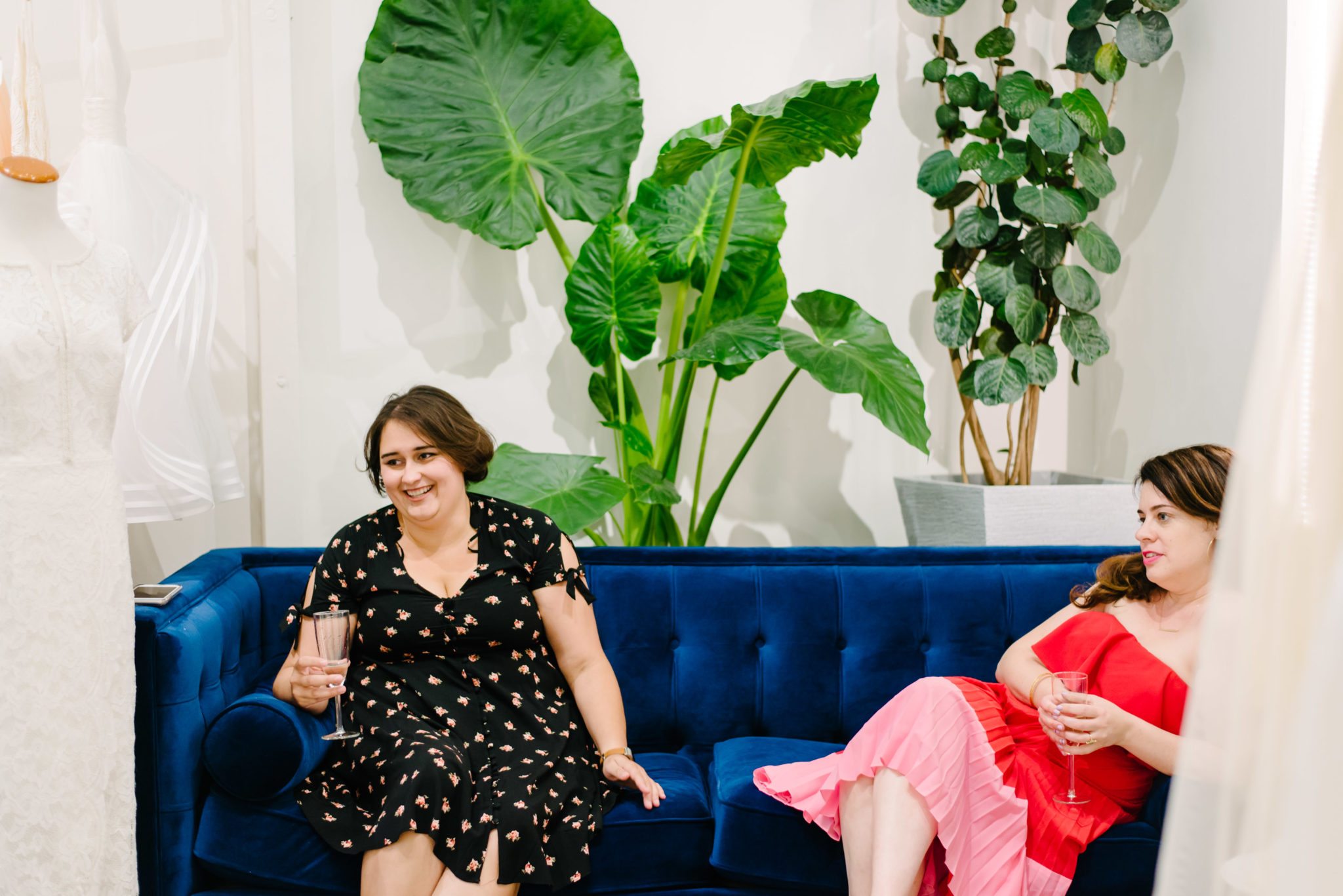 two women sit on a velvet couch, smiling, holding champagne glasses
