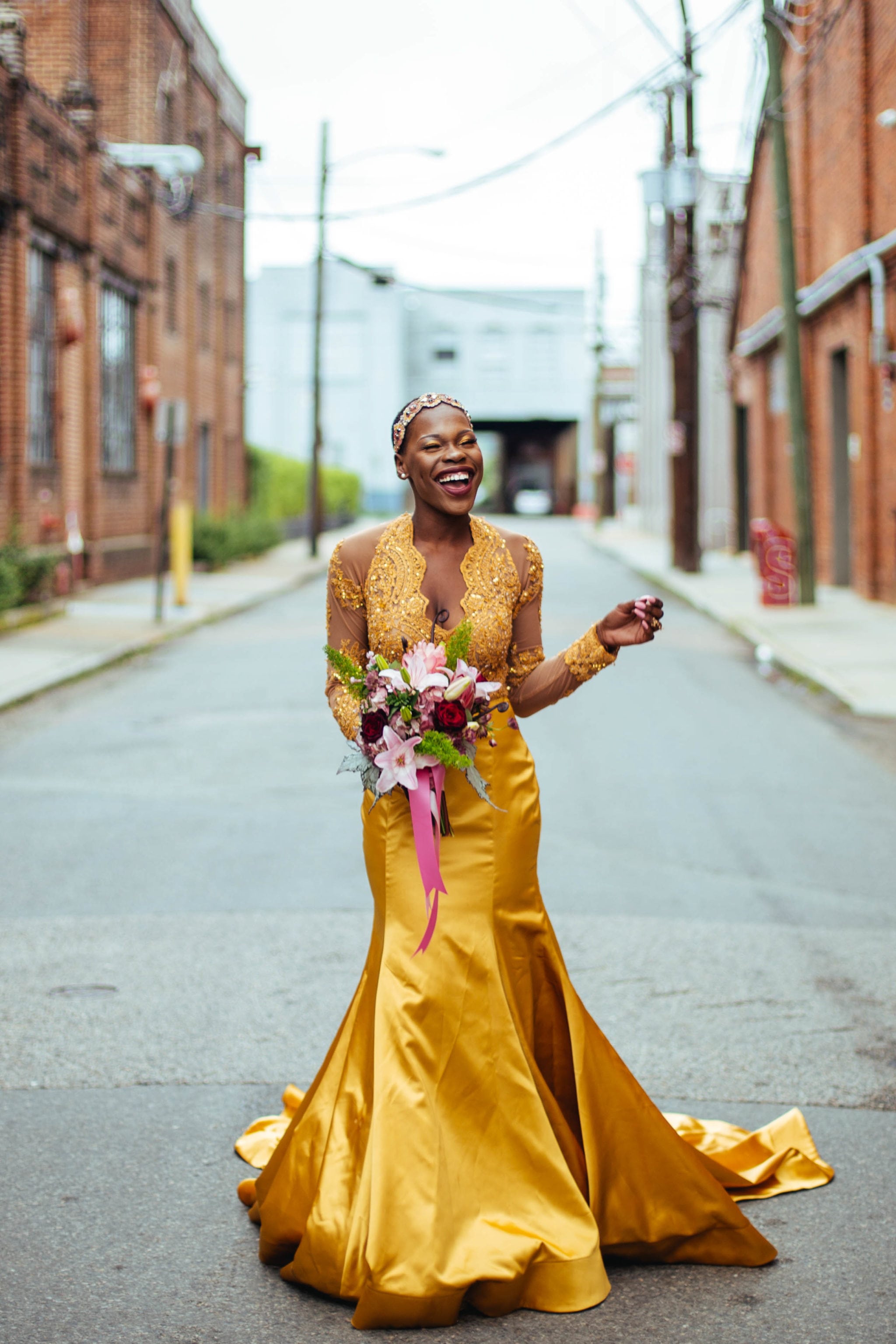 A strong woman stands in an alley wearing a golden dress, smiling, holding a bouquet