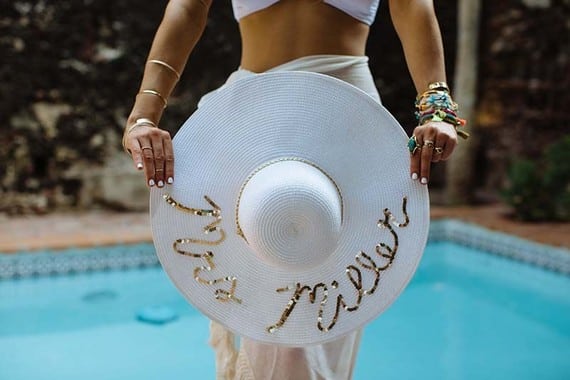 A woman in a white pool wrap with lots of bracelets stands in front of a pool holding a white sunhat with "Mrs. Miller" written in gold sequins on the brim.