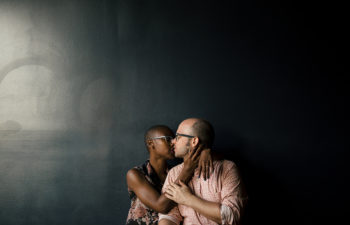 Couple wearing glasses embraces in front of a dark wall