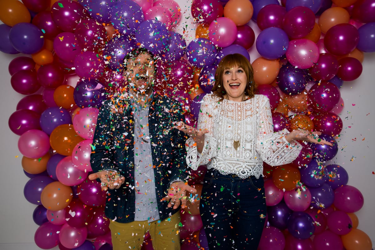 two people stand in front of a wall of balloons while being showered by confetti