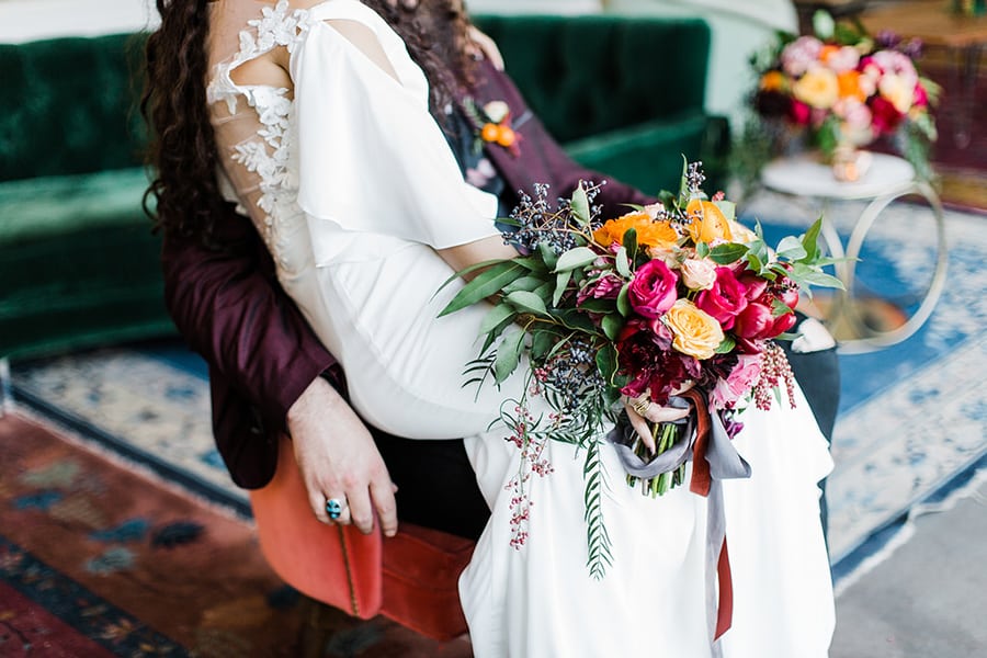 person in wedding dress holding colorful bouquet sits on partner's lap in an elegant indoor setting