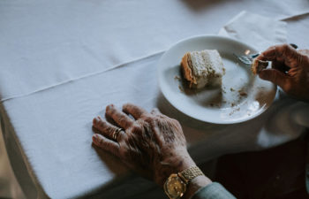An elderly hand wearing a large gold wristwatch rests on a white linen–clad table near a piece of cake on a white plate.