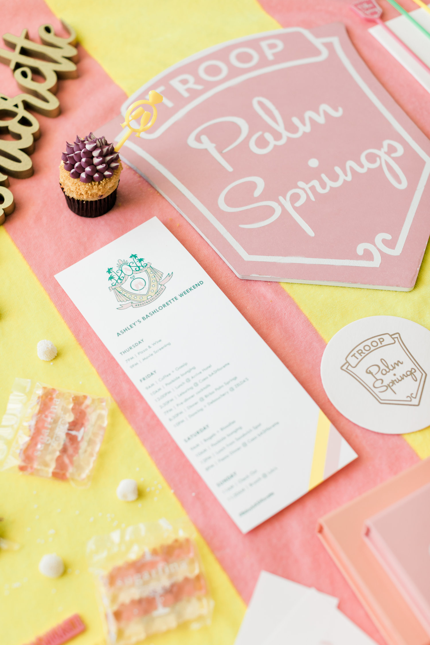 Troop Palm Springs pink, yellow, and white decor and printed collateral, including itinerary, mini cupcake, candies, and other bachelorette accoutrement