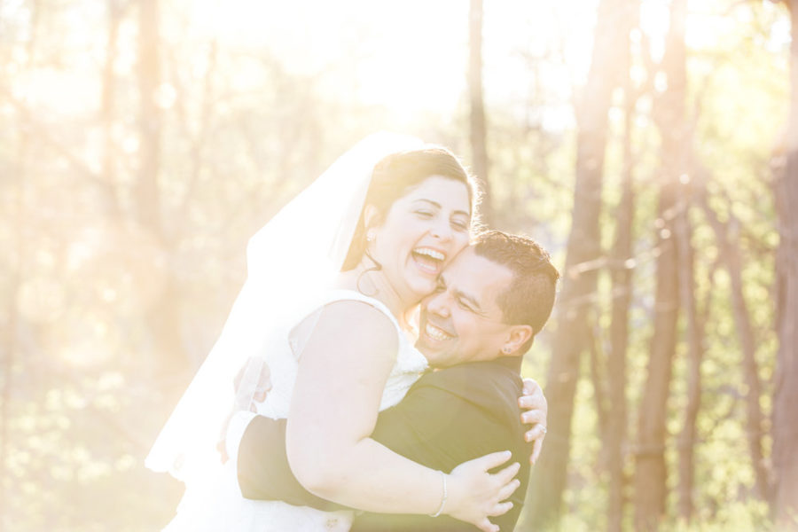 a wedding couple is lit from behind by the sun in the woods; both smile