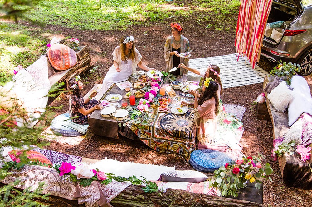 Five women wearing flower crowns casually lunch in a wooded area