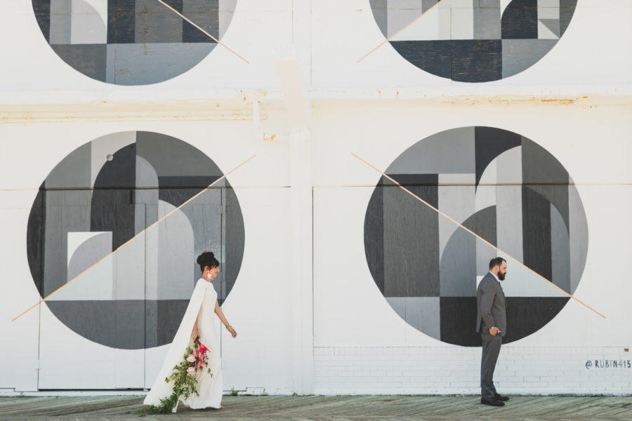 A woman with a floral whip bouquet wearing a white dress with a cape walks toward a man in a grey suit with his back to her as he stands in front of a white, grey, and black geometric wall
