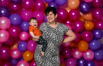 woman wearing a leopard print dress while holding a smiling baby in front of a wall of balloons