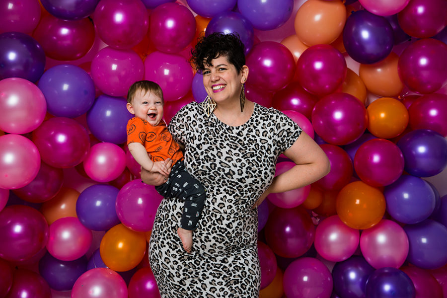 woman wearing a leopard print dress while holding a smiling baby in front of a wall of balloons