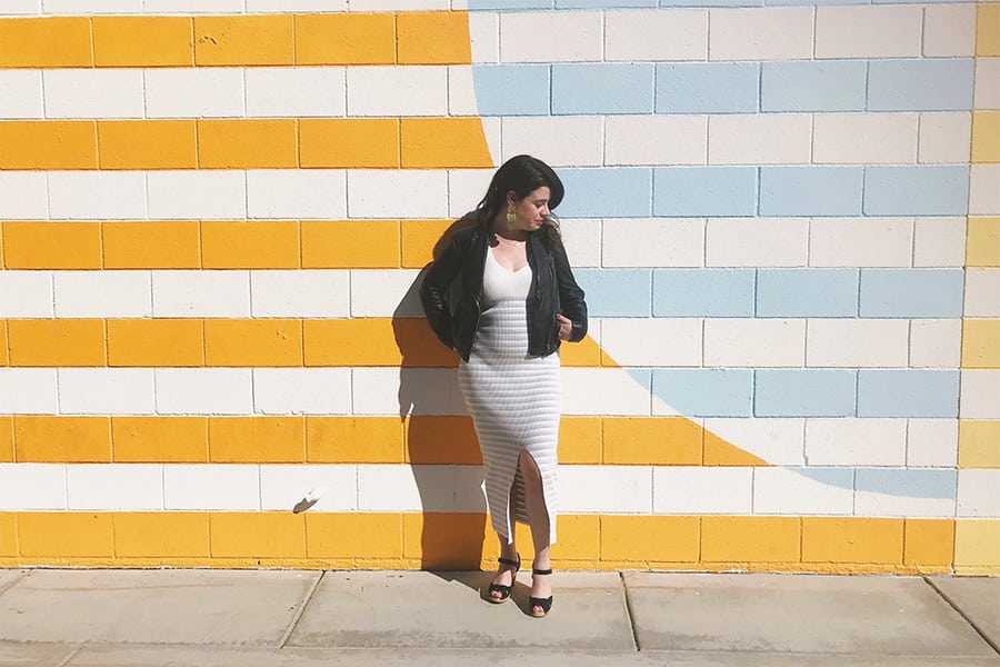 A woman leans against a yellow, white, and light blue striped wall wearing a leather jacket and white ankle-length sundress