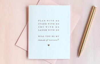 Notecard that reads: "Plan with me, stand with me, cry with me, laugh with me. Will you be my maid of honor?" on a pink envelope next to a gold pen.